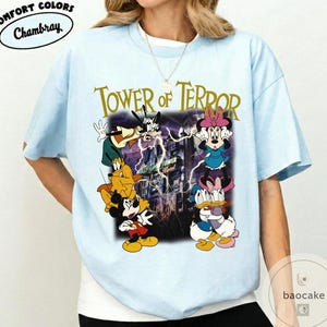 May include: Light blue t-shirt featuring a graphic of Disney characters, including Mickey Mouse, Goofy, and Minnie Mouse, with the text "TOWER OF TERROR" in gold lettering. The shirt has a crew neck and short sleeves. The brand name "Chambray" is in a white oval.