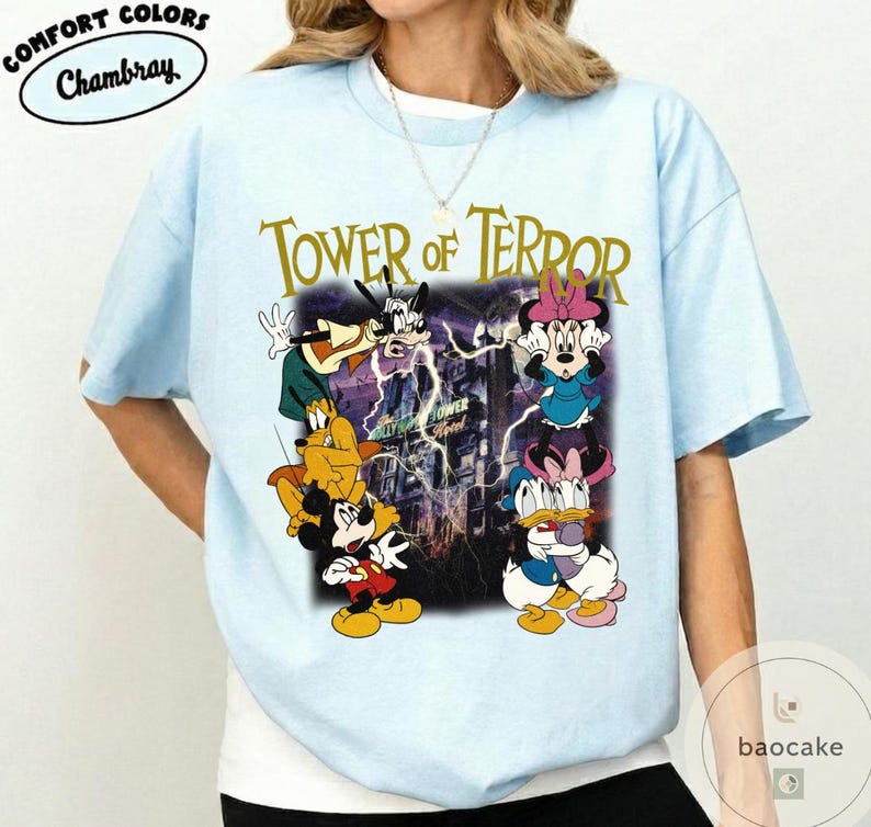 May include: Light blue t-shirt featuring a graphic of Disney characters, including Mickey Mouse, Goofy, and Minnie Mouse, with the text "TOWER OF TERROR" in gold lettering. The shirt has a crew neck and short sleeves. The brand name "Chambray" is in a white oval.