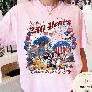Puede incluir: Camiseta rosa claro con un gr&aacute;fico colorido que celebra 250 a&ntilde;os. El dise&ntilde;o incluye a Mickey Mouse, Minnie Mouse, Donald Duck y Pluto, junto con banderas americanas, fuegos artificiales y globos. El texto incluye "Oh Boy!" y "Celebrating Of Joy."