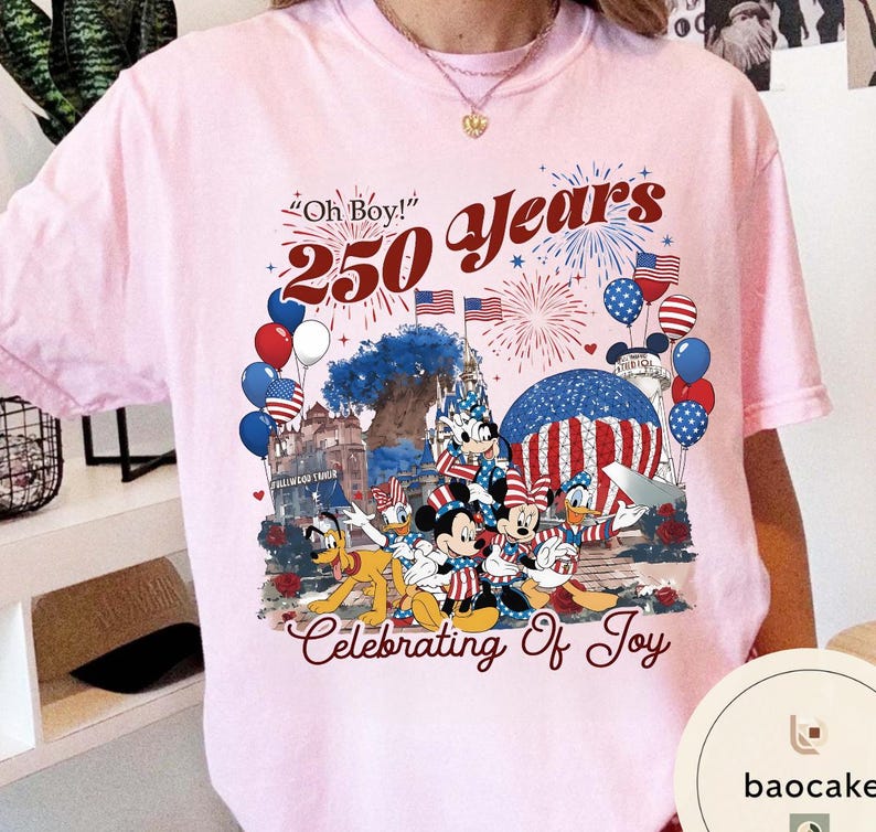 Puede incluir: Camiseta rosa claro con un gr&aacute;fico colorido que celebra 250 a&ntilde;os. El dise&ntilde;o incluye a Mickey Mouse, Minnie Mouse, Donald Duck y Pluto, junto con banderas americanas, fuegos artificiales y globos. El texto incluye "Oh Boy!" y "Celebrating Of Joy."