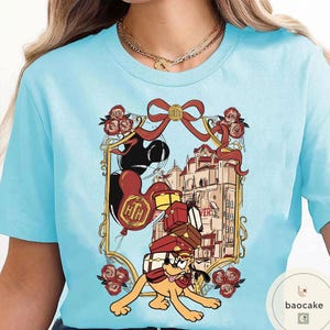 May include: Light blue t-shirt featuring a cartoon illustration. The design includes a dog carrying luggage, balloons, and a building. The graphic is framed with a red ribbon and roses. The shirt has a round neckline and short sleeves.