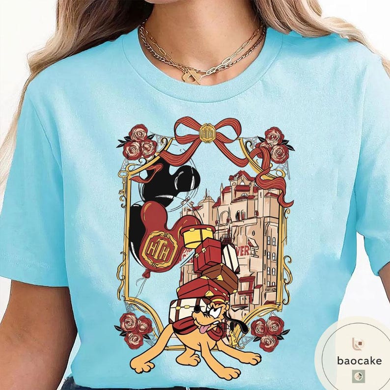 May include: Light blue t-shirt featuring a cartoon illustration. The design includes a dog carrying luggage, balloons, and a building. The graphic is framed with a red ribbon and roses. The shirt has a round neckline and short sleeves.