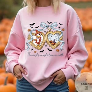 May include: Pale pink sweatshirt with a Halloween-themed heart-shaped locket design. The locket features cartoon characters and spooky imagery. The text "The most scariest place on earth" is printed below. Bats, spiders, and pumpkins are scattered around.