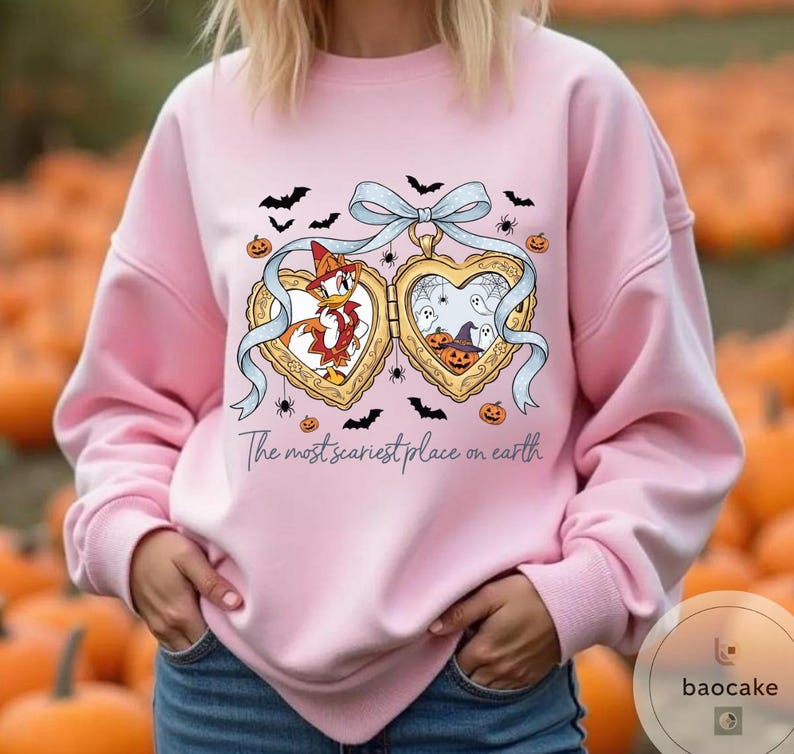 May include: Pale pink sweatshirt with a Halloween-themed heart-shaped locket design. The locket features cartoon characters and spooky imagery. The text "The most scariest place on earth" is printed below. Bats, spiders, and pumpkins are scattered around.