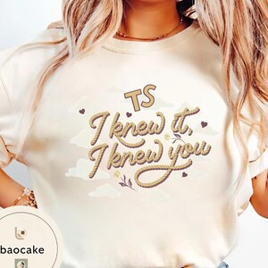 K&ouml;nnte beinhalten: Ein cremefarbenes Sweatshirt mit Rundhalsausschnitt und einem Grafikdesign mit dem Text TS I knew it, I knew you in einer seilartigen Schriftart. Das Design enth&auml;lt kleine braune Herzen, hellbeige Wolkenformen und florale Linienzeichnungen.
