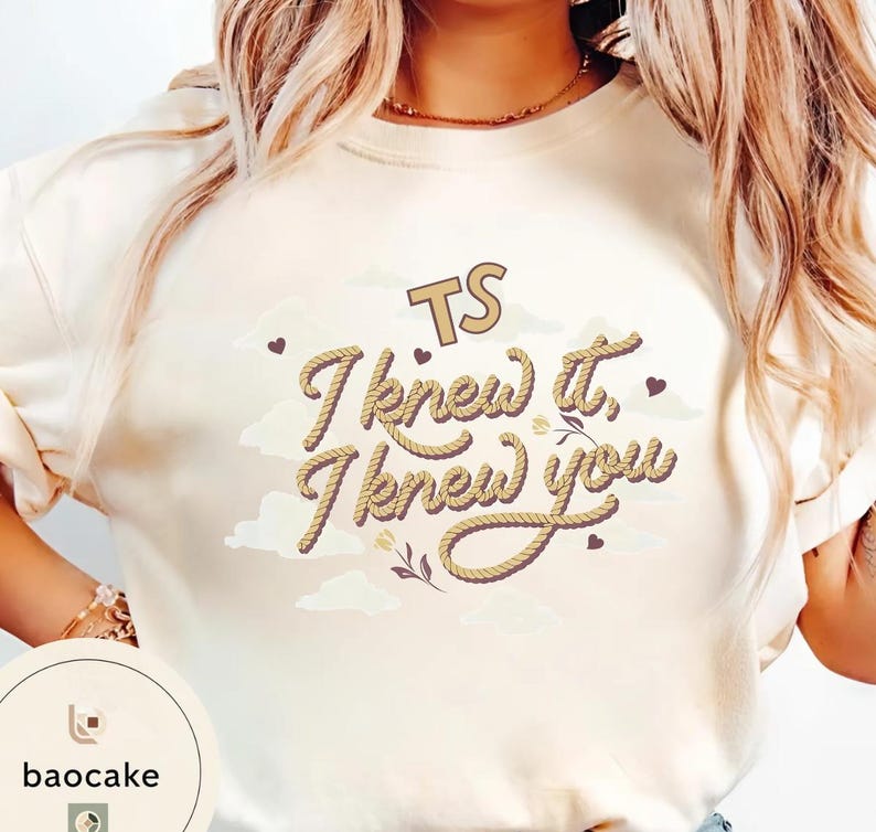 K&ouml;nnte beinhalten: Ein cremefarbenes Sweatshirt mit Rundhalsausschnitt und einem Grafikdesign mit dem Text TS I knew it, I knew you in einer seilartigen Schriftart. Das Design enth&auml;lt kleine braune Herzen, hellbeige Wolkenformen und florale Linienzeichnungen.
