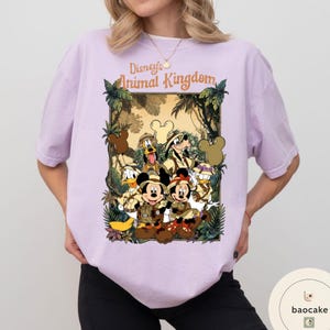 May include: A light purple t-shirt featuring a graphic of Disney's Animal Kingdom characters, including Mickey and Minnie Mouse, Donald Duck, and Goofy, in safari attire. The text "Disney's Animal Kingdom" is arched above the characters.