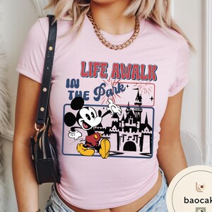 May include: Pale pink t-shirt with a graphic of Mickey Mouse and a castle, with the text "LIFE A WALK IN THE PARK". The design includes fireworks and a blue border. The shirt has short sleeves.