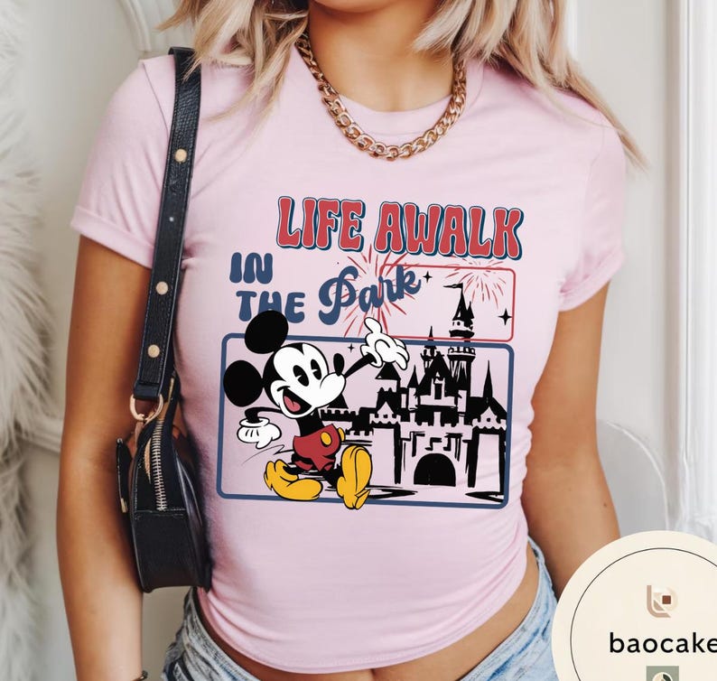 May include: Pale pink t-shirt with a graphic of Mickey Mouse and a castle, with the text "LIFE A WALK IN THE PARK". The design includes fireworks and a blue border. The shirt has short sleeves.