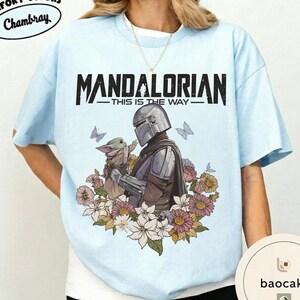 Puede incluir: Camiseta azul claro con el texto negro "MANDALORIAN - THIS IS THE WAY". El dise&ntilde;o muestra un personaje mandaloriano sosteniendo una peque&ntilde;a criatura, rodeado de flores y mariposas.