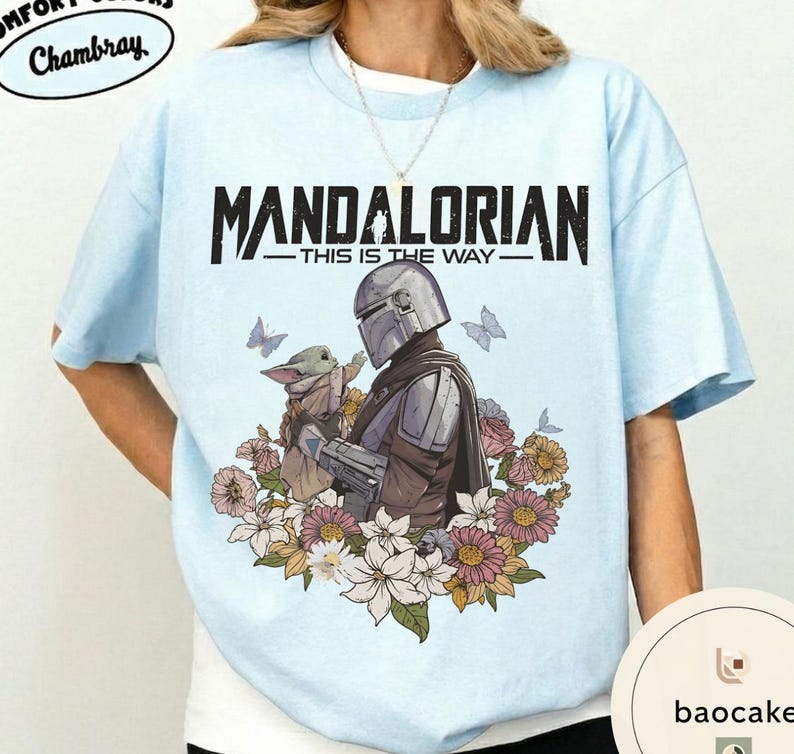 Puede incluir: Camiseta azul claro con el texto negro "MANDALORIAN - THIS IS THE WAY". El dise&ntilde;o muestra un personaje mandaloriano sosteniendo una peque&ntilde;a criatura, rodeado de flores y mariposas.