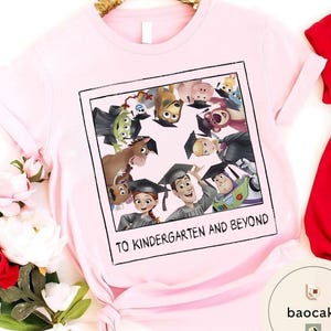 May include: A pale pink t-shirt with a graphic of Toy Story characters in graduation attire, set within a polaroid frame. The text "TO KINDERGARTEN AND BEYOND" is printed below. The shirt has short sleeves and is made of a soft material.