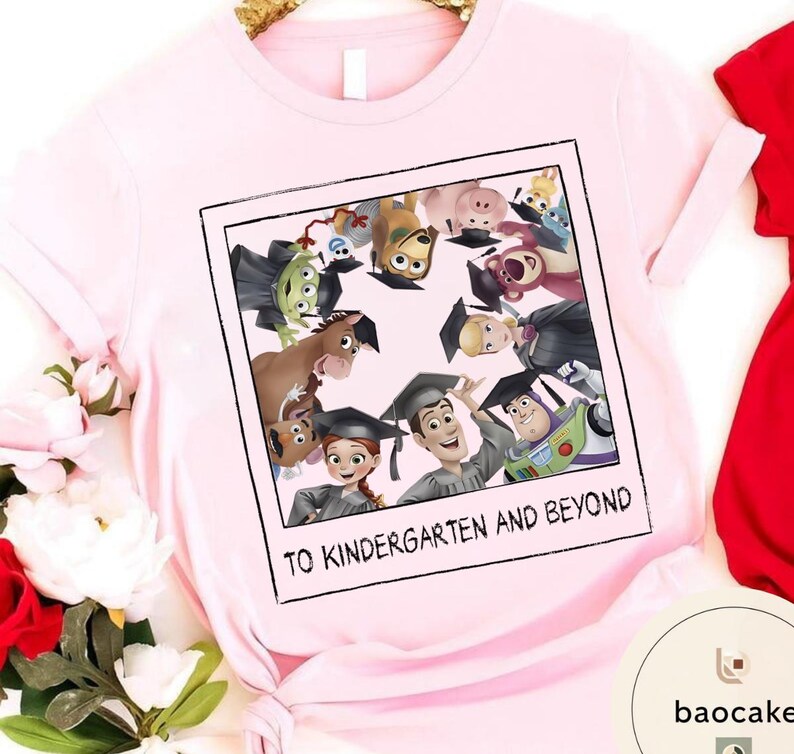 May include: A pale pink t-shirt with a graphic of Toy Story characters in graduation attire, set within a polaroid frame. The text "TO KINDERGARTEN AND BEYOND" is printed below. The shirt has short sleeves and is made of a soft material.