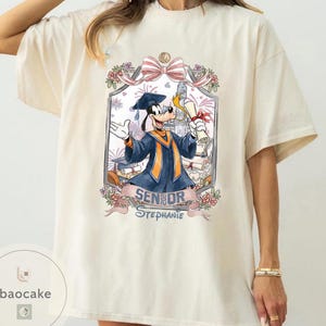 May include: Cream-colored t-shirt featuring a graduation-themed graphic. The graphic includes a cartoon character in a graduation gown, a diploma, and the text "SENIOR Stephanie". The design is framed with floral and bow accents.