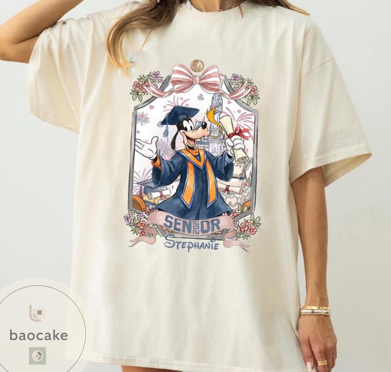 May include: Cream-colored t-shirt featuring a graduation-themed graphic. The graphic includes a cartoon character in a graduation gown, a diploma, and the text "SENIOR Stephanie". The design is framed with floral and bow accents.