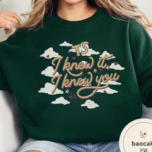 K&ouml;nnte beinhalten: Ein dunkelgr&uuml;ner Rundhals-Pullover mit einem Grafikdruck aus wei&szlig;en Wolken, kleinen violetten Herzen und zwei kleinen Blumen. Der Text "TS I knew it, I knew you" ist in einer beigen, seilartigen Schriftart auf der Vorderseite abgebildet.