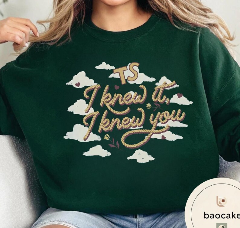 K&ouml;nnte beinhalten: Ein dunkelgr&uuml;ner Rundhals-Pullover mit einem Grafikdruck aus wei&szlig;en Wolken, kleinen violetten Herzen und zwei kleinen Blumen. Der Text "TS I knew it, I knew you" ist in einer beigen, seilartigen Schriftart auf der Vorderseite abgebildet.