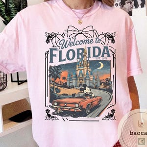 May include: Pink t-shirt featuring a graphic with the text "Welcome to Florida" and a vintage-style illustration. The illustration includes a castle, palm trees, a car with Mickey Mouse, and a crescent moon.