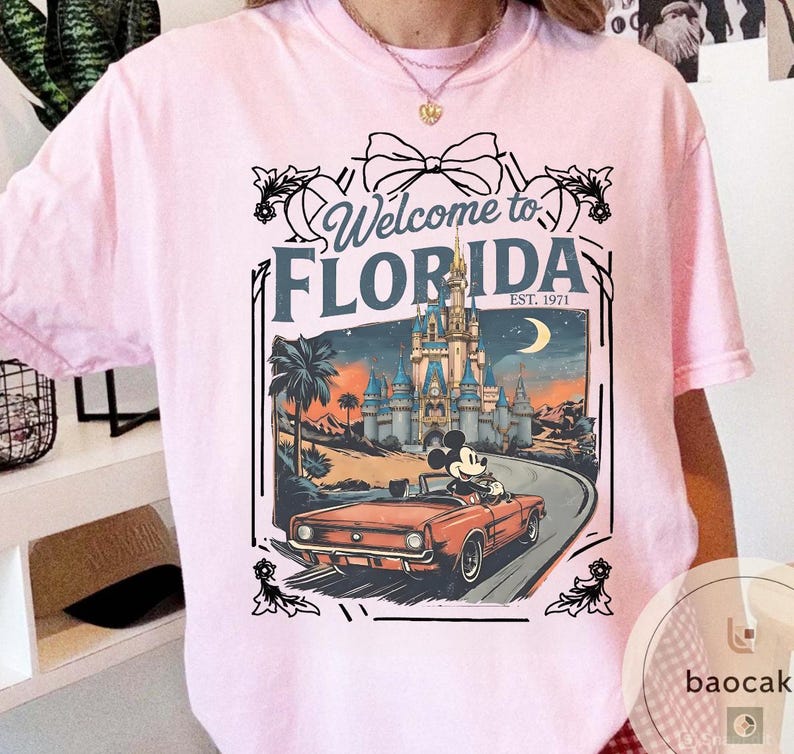 May include: Pink t-shirt featuring a graphic with the text "Welcome to Florida" and a vintage-style illustration. The illustration includes a castle, palm trees, a car with Mickey Mouse, and a crescent moon.