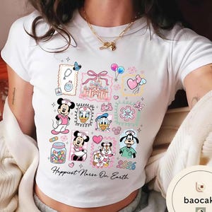 May include: White t-shirt featuring colorful cartoon illustrations of Disney characters in nurse attire, including Mickey and Minnie Mouse, Donald Duck, and Goofy. The shirt includes the text "Happiest Nurse On Earth" and the year "2026".