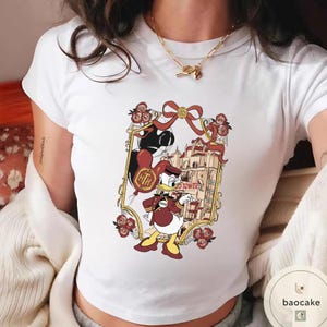 May include: White t-shirt featuring a cartoon graphic of Donald Duck in a maroon and gold outfit, holding a balloon and a sign that reads "TD Tower". The graphic is framed with roses and a red bow.