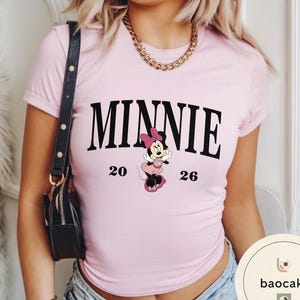 May include: A light pink t-shirt featuring the word "MINNIE" in bold black letters. Below the word is a cartoon image of Minnie Mouse in a pink dress and bow. The numbers "20" and "26" are printed on either side of the character.