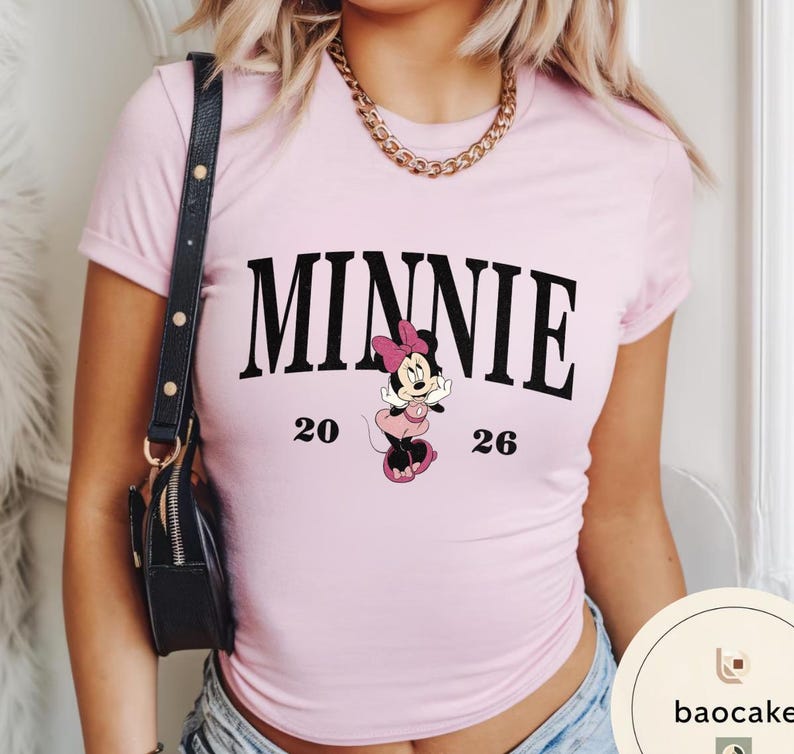 May include: A light pink t-shirt featuring the word "MINNIE" in bold black letters. Below the word is a cartoon image of Minnie Mouse in a pink dress and bow. The numbers "20" and "26" are printed on either side of the character.