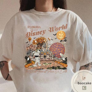 Op de afbeelding: Cr&egrave;mekleurig T-shirt met een Halloween-thema van Disney World. Het ontwerp bevat een mummie, pompoenen, een kasteel en de tekst "Florida Disney World Est 2018" en "The Most Scariest Place On Earth."