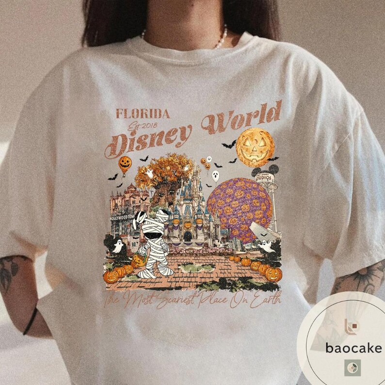 Op de afbeelding: Cr&egrave;mekleurig T-shirt met een Halloween-thema van Disney World. Het ontwerp bevat een mummie, pompoenen, een kasteel en de tekst "Florida Disney World Est 2018" en "The Most Scariest Place On Earth."