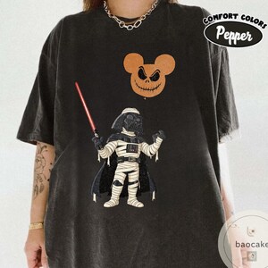 May include: Black t-shirt featuring a cartoon illustration of Darth Vader holding a lightsaber, with a Mickey Mouse-shaped Jack Skellington balloon. The shirt has the words "Comfort Colors Pepper" printed on it.