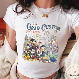 May include: White t-shirt with a colourful graphic of Disney characters in graduation attire. The design includes the text "Grad Custom Class of 2026" with a castle and fireworks. A graduation cap and books are also visible.