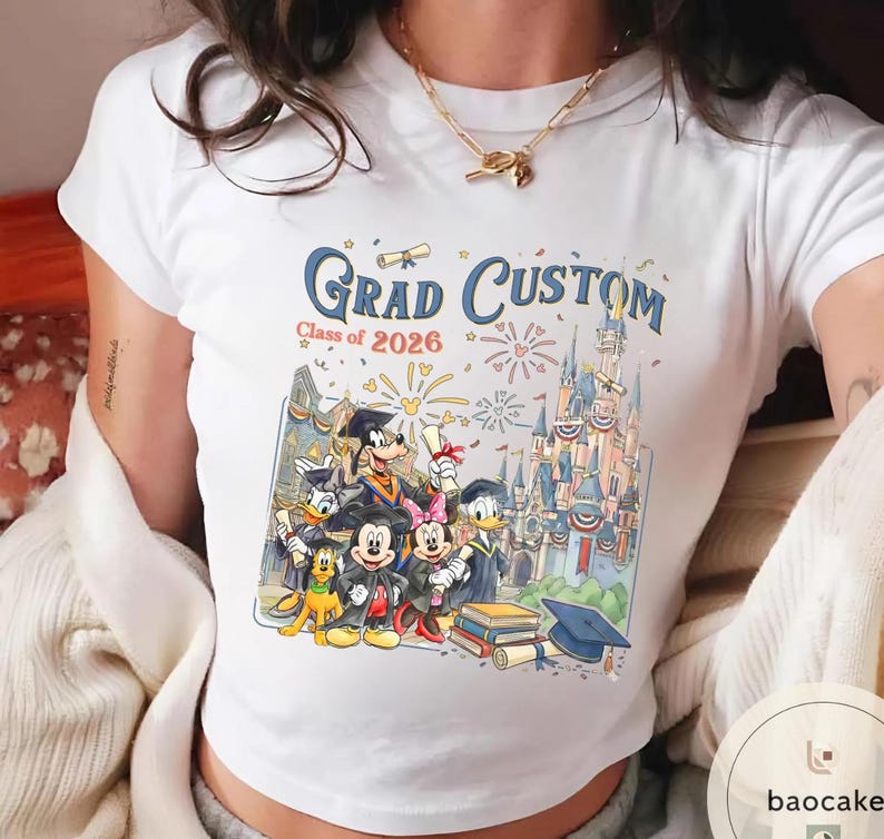 May include: White t-shirt with a colourful graphic of Disney characters in graduation attire. The design includes the text "Grad Custom Class of 2026" with a castle and fireworks. A graduation cap and books are also visible.