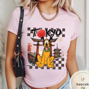 Puede incluir: Camiseta rosa claro con un dise&ntilde;o de Pluto frente a un templo japon&eacute;s con la palabra "TOKYO" encima. El dise&ntilde;o incluye un sol rojo, fuegos artificiales y un patr&oacute;n de tablero de ajedrez. La camiseta tiene mangas cortas.