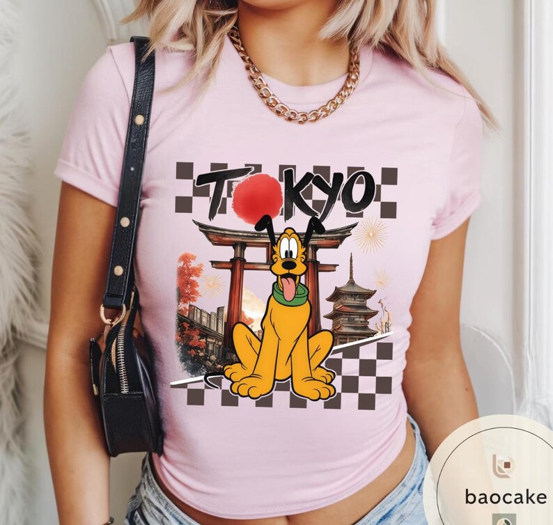 Puede incluir: Camiseta rosa claro con un dise&ntilde;o de Pluto frente a un templo japon&eacute;s con la palabra "TOKYO" encima. El dise&ntilde;o incluye un sol rojo, fuegos artificiales y un patr&oacute;n de tablero de ajedrez. La camiseta tiene mangas cortas.