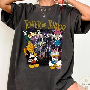 May include: Dark gray t-shirt featuring the text "Tower of Terror" above a graphic of cartoon characters including Mickey Mouse, Minnie Mouse, Donald Duck, and Goofy. The graphic has a purple and yellow color scheme.