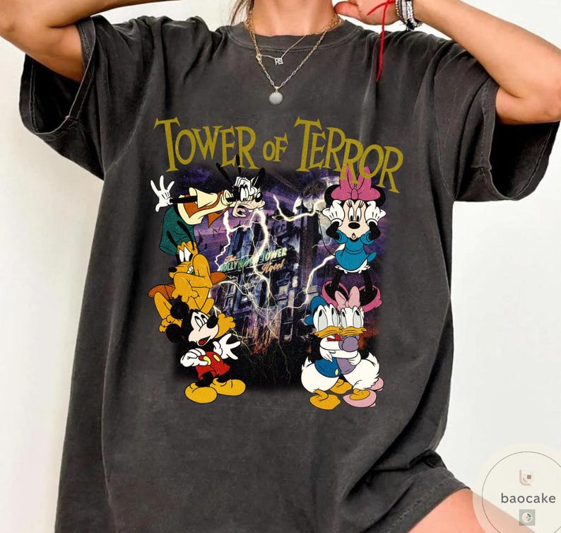 May include: Dark gray t-shirt featuring the text "Tower of Terror" above a graphic of cartoon characters including Mickey Mouse, Minnie Mouse, Donald Duck, and Goofy. The graphic has a purple and yellow color scheme.