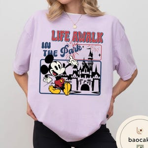 May include: A lilac t-shirt with a graphic of Mickey Mouse gesturing towards a castle. The text "LIFE A WALK IN THE Park" is above the graphic in red and blue. The shirt has short sleeves and a crew neck.