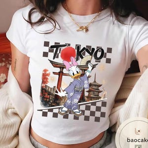 Puede incluir: Camiseta blanca con un dise&ntilde;o de Daisy Duck con kimono, sosteniendo un abanico, con la palabra "TOKYO" encima. El dise&ntilde;o incluye una pagoda japonesa y un patr&oacute;n de tablero de ajedrez.