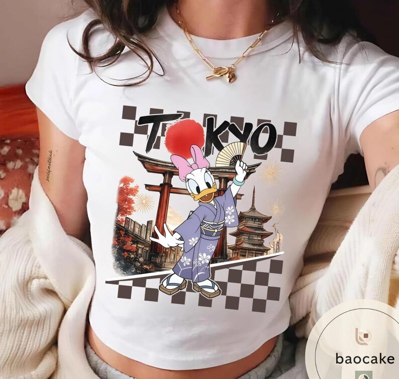 Puede incluir: Camiseta blanca con un dise&ntilde;o de Daisy Duck con kimono, sosteniendo un abanico, con la palabra "TOKYO" encima. El dise&ntilde;o incluye una pagoda japonesa y un patr&oacute;n de tablero de ajedrez.