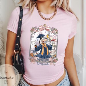 May include: Light pink t-shirt featuring a cartoon illustration of Goofy in a graduation gown, holding a diploma. The design includes the text "Senior Stephanie" and floral accents. The shirt has short sleeves and a crew neck.
