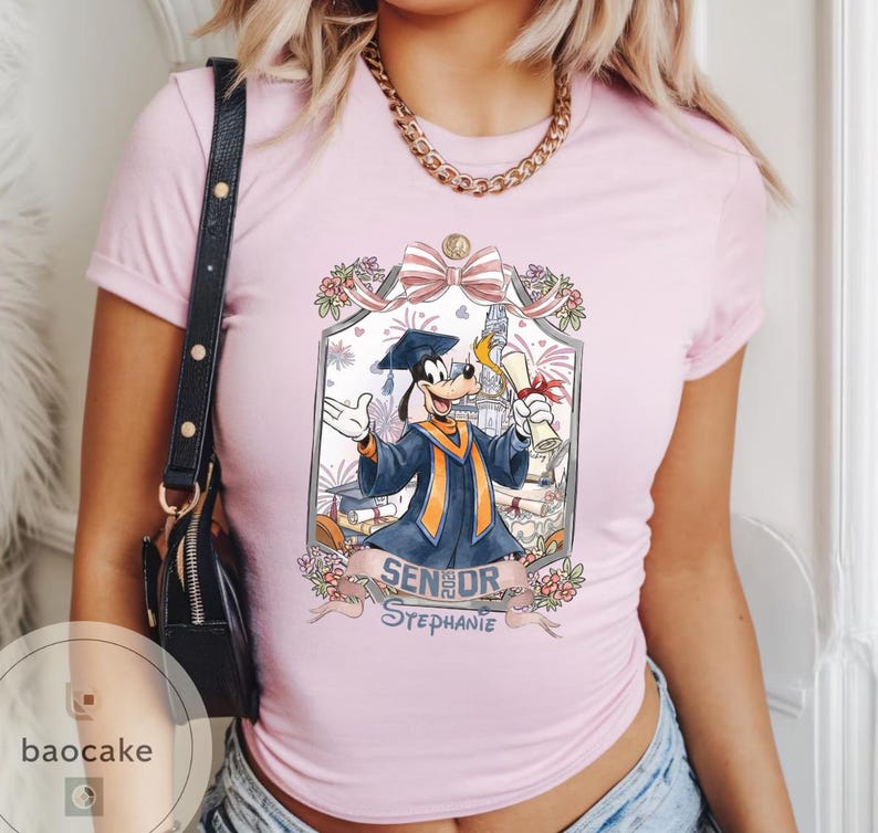 May include: Light pink t-shirt featuring a cartoon illustration of Goofy in a graduation gown, holding a diploma. The design includes the text "Senior Stephanie" and floral accents. The shirt has short sleeves and a crew neck.