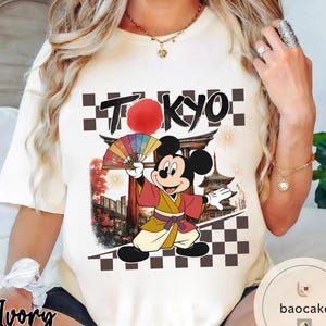 Puede incluir: Camiseta color crema con un dise&ntilde;o de Mickey Mouse con ropa japonesa, sosteniendo un abanico. La palabra "TOKYO" est&aacute; encima del personaje, con un sol rojo y detalles a cuadros. El dise&ntilde;o incluye arquitectura japonesa y fuegos artificiales.