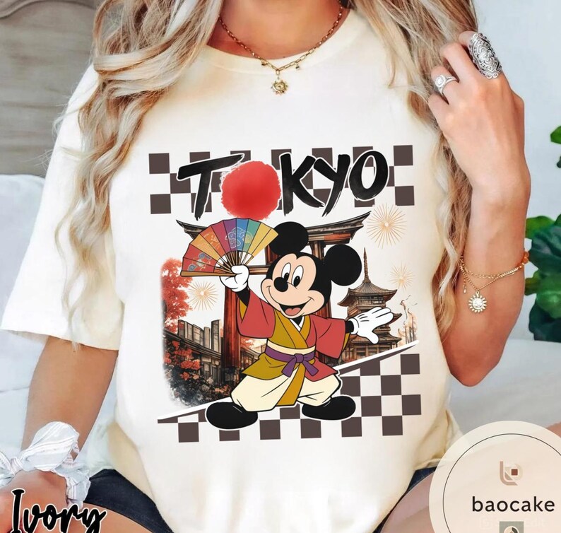 Puede incluir: Camiseta color crema con un dise&ntilde;o de Mickey Mouse con ropa japonesa, sosteniendo un abanico. La palabra "TOKYO" est&aacute; encima del personaje, con un sol rojo y detalles a cuadros. El dise&ntilde;o incluye arquitectura japonesa y fuegos artificiales.