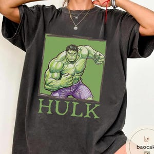 May include: Dark gray t-shirt featuring a green and purple illustration of the Hulk character. The illustration is set against a green background with the word "HULK" in green block letters below the image. The shirt has short sleeves.