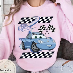 May include: A light pink sweatshirt featuring a blue cartoon car with green eyes and a smiling face. The design includes checkered flags and racing stripes. The sweatshirt has a crew neck and long sleeves.