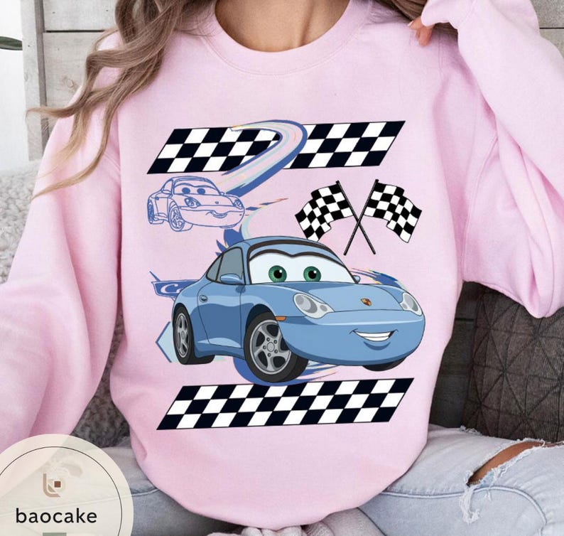 May include: A light pink sweatshirt featuring a blue cartoon car with green eyes and a smiling face. The design includes checkered flags and racing stripes. The sweatshirt has a crew neck and long sleeves.