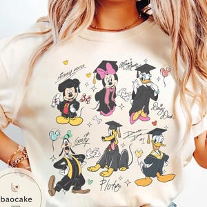 May include: Cream-colored t-shirt featuring cartoon characters Mickey Mouse, Minnie Mouse, Donald Duck, Daisy Duck, Goofy, and Pluto in graduation attire. Each character is wearing a graduation cap and gown. The shirt includes character names and signatures.
