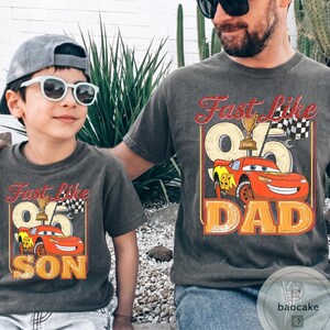 May include: Two matching charcoal gray t-shirts. One says "Fast Like 95 SON" with a cartoon race car. The other says "Fast Like 95 DAD" with the same car graphic. Both shirts have a vintage look.