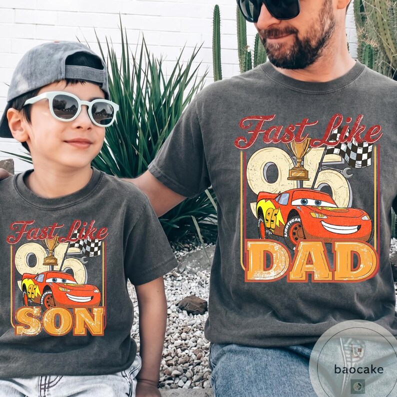 May include: Two matching charcoal gray t-shirts. One says "Fast Like 95 SON" with a cartoon race car. The other says "Fast Like 95 DAD" with the same car graphic. Both shirts have a vintage look.