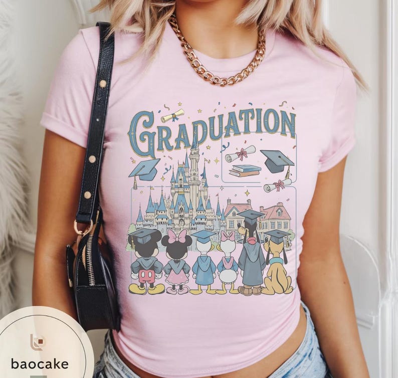 May include: A light pink t-shirt featuring a graphic with the word "Graduation" in a vintage font. The graphic includes a castle, graduation caps, and cartoon characters like Mickey and Minnie Mouse.